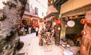 A vibrant outdoor market scene displaying hanging brass pots and people shopping.