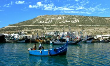 Fishing boats anchored in Agadir port under a hill with a prominent Arabic inscription.
