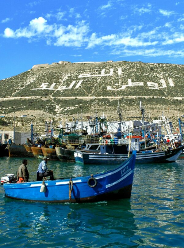 Fishing boats anchored in Agadir port under a hill with a prominent Arabic inscription.