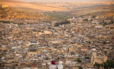 From above back view of unrecognizable female friends in traditional wear sitting on hilltop and looking at town with typical buildings