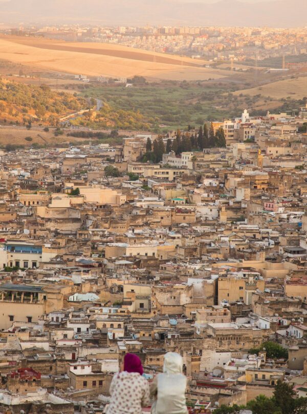 From above back view of unrecognizable female friends in traditional wear sitting on hilltop and looking at town with typical buildings